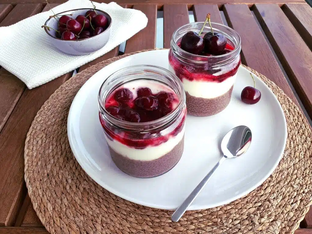 Zwei Gläser Schoko Chia Pudding mit Joghurt und Kirschen auf einem weißen Teller, der auf einem Holztisch steht.