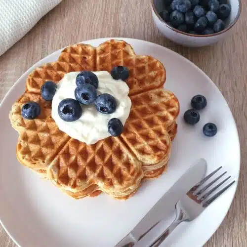 Ein Stapel High Protein Waffeln auf einem weißen Teller, serviert mit Blaubeeren und Quark, mit Besteck und einer Schale Beeren auf einem Tisch.